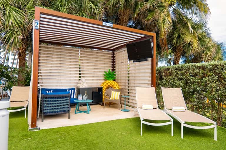 Beachside cabana with wood frame, louvered ceiling, ocean view, blue seating, and adjacent loungers on grass