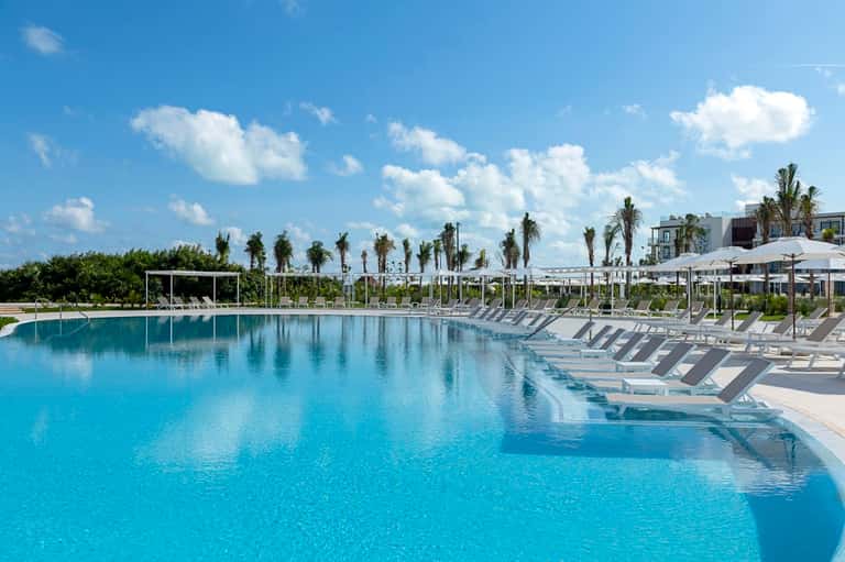 Large resort swimming pool with crystal-clear turquoise water, lounge chairs, umbrellas, and palm tree landscaping