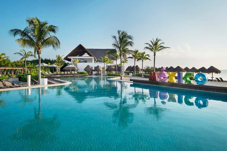 Modern resort pool with turquoise water, white buildings, palm trees, and colorful letters spelling resort name