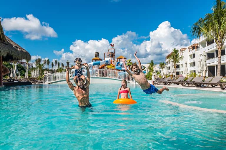 Family enjoying shallow pool area with water slide and colorful umbrellas at resort