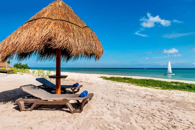 Tropical beach with thatched umbrella, lounge chair, sailboat, and clear turquoise water