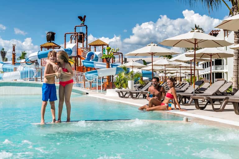 Family-friendly resort pool area with water slide, families swimming, umbrellas, and white buildings