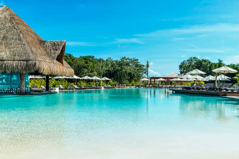 Tropical resort lagoon pool with thatch-roof bar, white umbrellas, loungers, and palm trees