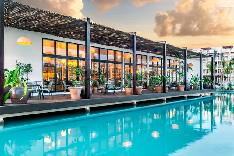 Modern poolside restaurant with large windows, lounge seating, and turquoise pool at dusk