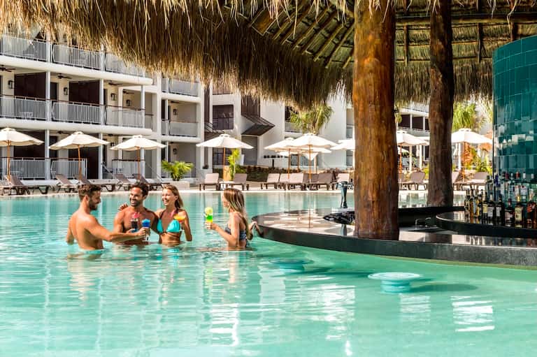 Guests enjoying cocktails in turquoise pool beneath thatched structure with resort buildings and umbrellas