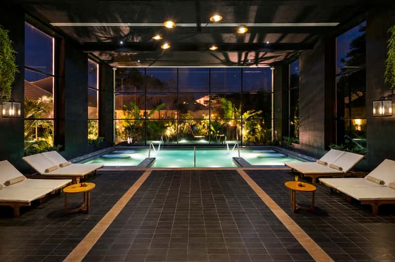 Contemporary indoor pool with glass walls, lounge chairs, ambient lighting, and evening ambiance