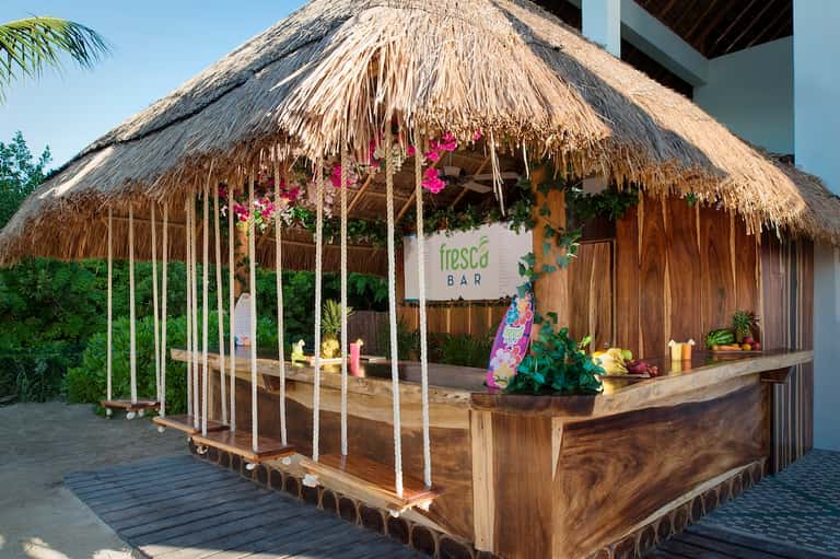 Tropical bar structure with thatched roof, wooden counter, pink flowers, and palm vegetation