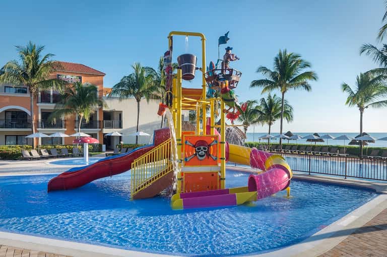 Children's water play area with colorful slide structure in resort pool surrounded by palm trees