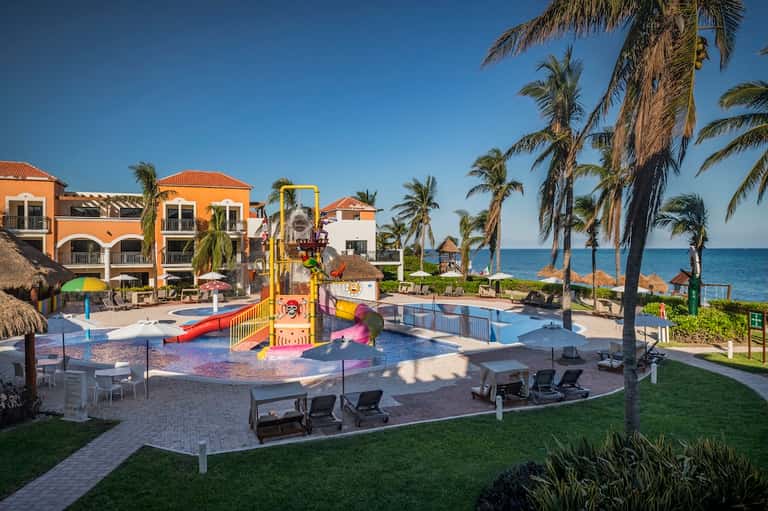 Beachfront resort with colorful buildings, pool, playground, palm trees, and ocean view
