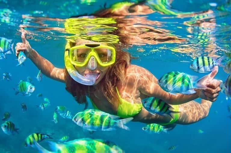 Snorkeler underwater smiling at camera with colorful tropical fish swimming nearby in clear blue water