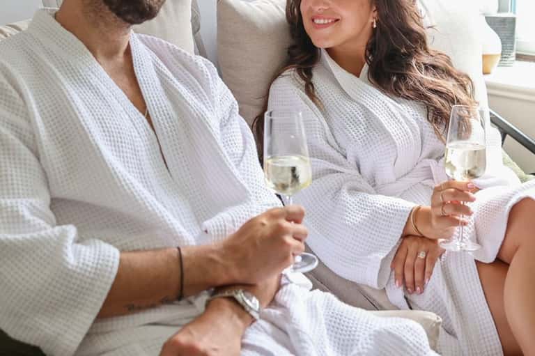 Couple in white robes toasting with champagne glasses indoors