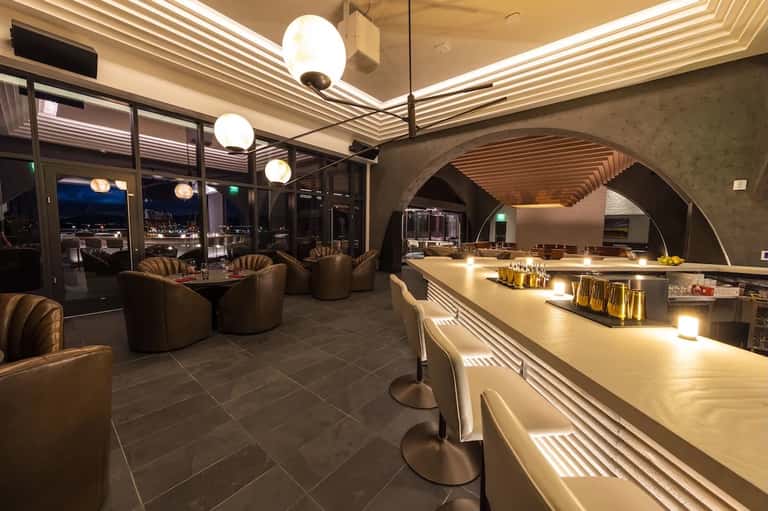 Modern lounge bar interior at night with ambient lighting, curved seating, and city views through floor-to-ceiling windows