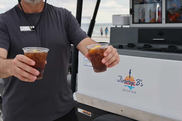 Beach bar server holding two cola drinks with ocean and beachgoers in background