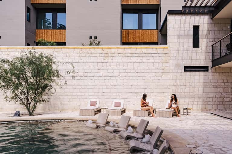 Two women relaxing by courtyard pool with concrete loungers and stone building walls