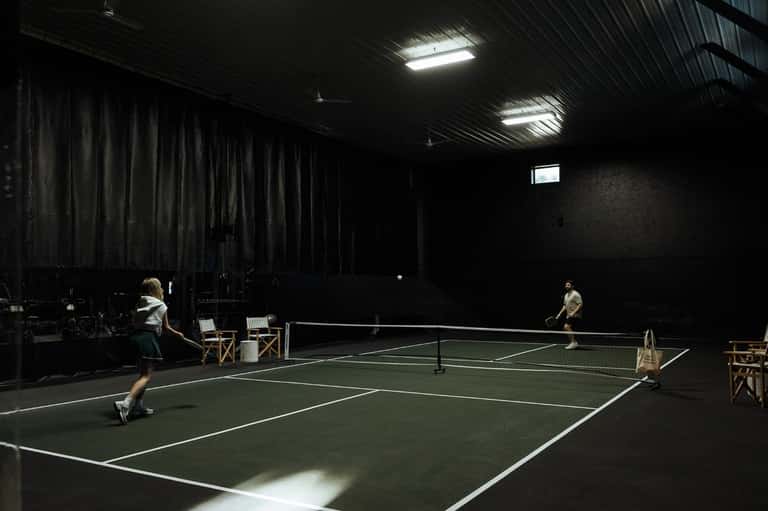 Indoor pickleball court with players, fluorescent lighting, and spectator seating