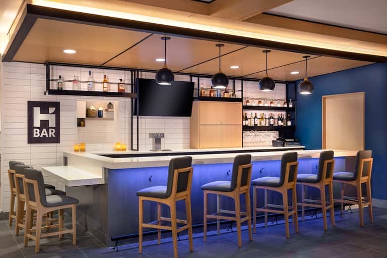Modern hotel bar with blue lighting, wooden bar stools, and shelved liquor display