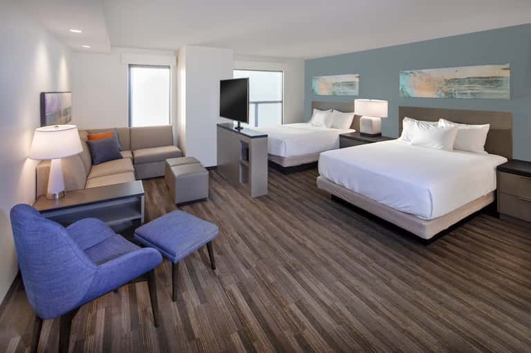Modern hotel suite with two beds, lounge area, blue accent wall, and contemporary furnishings
