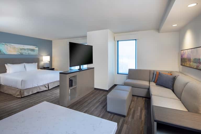 Modern hotel suite with king bed, TV, gray sofa seating area, and contemporary furnishings