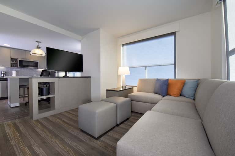 Modern hotel suite with gray sofa, kitchen area, TV, and large bright windows