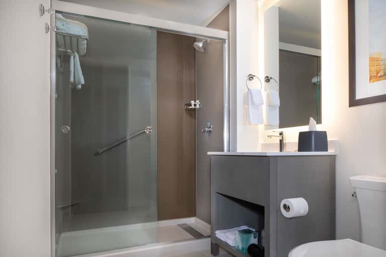 Modern bathroom with glass shower enclosure, gray vanity with white sink, white towels, and contemporary fixtures.
