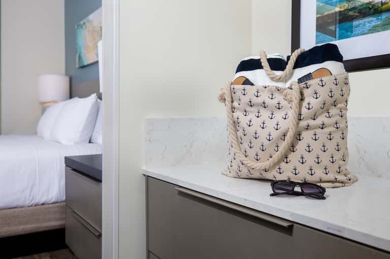 Bathroom vanity with anchor-patterned tote bag and sunglasses, bedroom visible through glass door