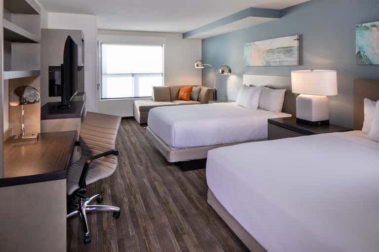 Modern hotel room with two beds, work desk, orange accent pillow, and blue wall accents