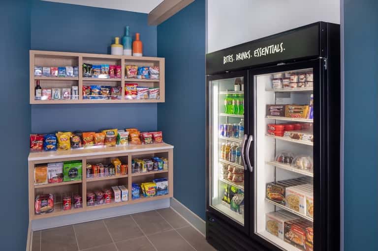 Hotel snack station with wooden shelving, vending refrigerator, and colorful packaged snacks and beverages.