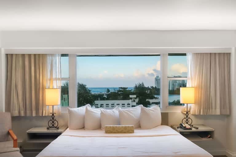 Hotel bedroom with ocean view windows, white bedding, elegant lamps, and coastal scenery
