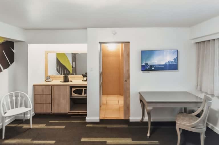 Hotel room kitchenette and living area with wood cabinetry and glass dining table