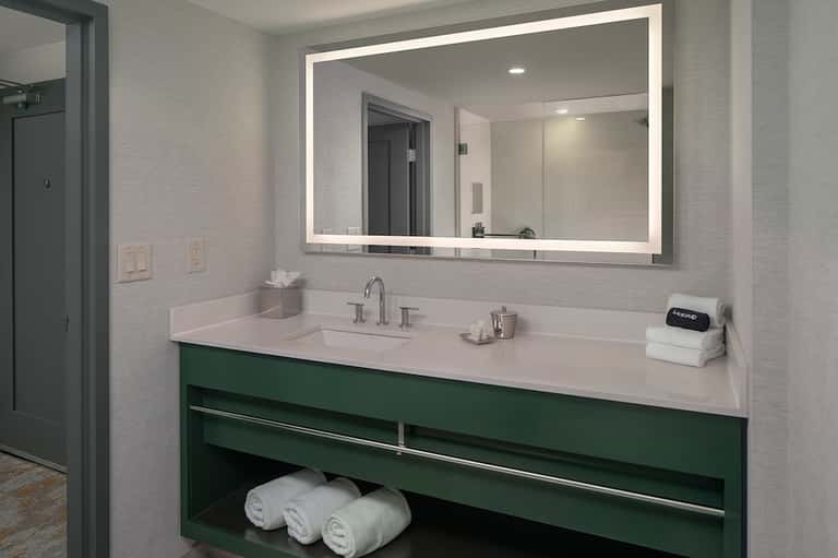 Modern hotel bathroom with illuminated mirror, green vanity cabinet, white countertop, and folded towels