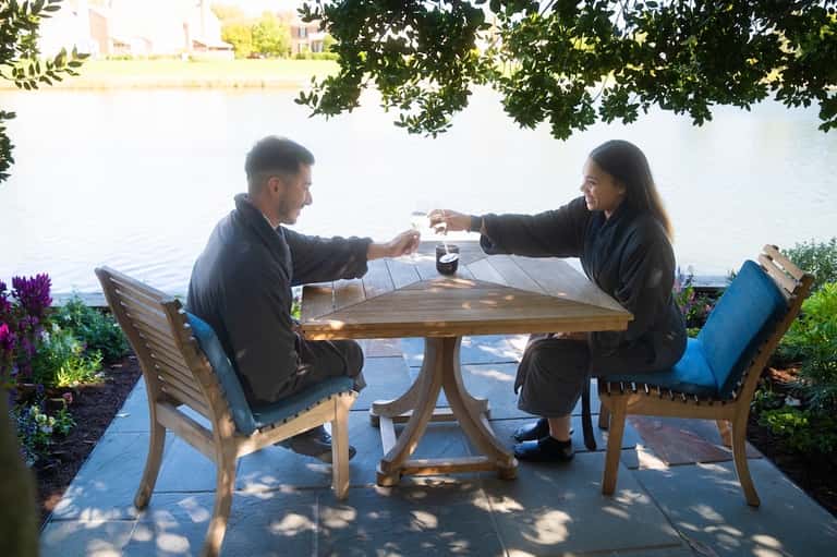 Couple toasting drinks at waterside table with tree shade and scenic water backdrop