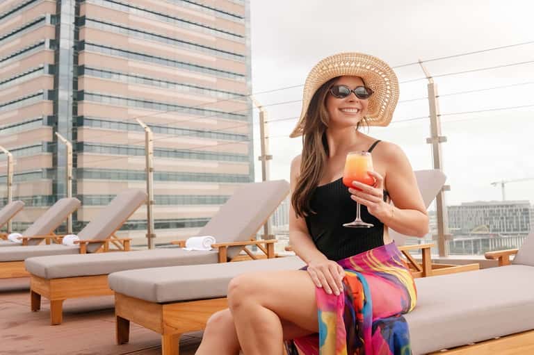 Woman in sun hat enjoying cocktail on rooftop lounge with city skyline and wooden deck chairs visible