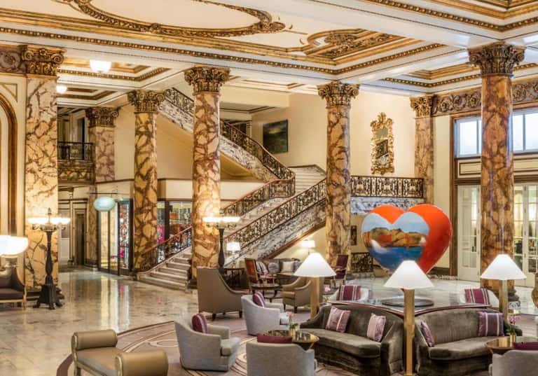 Grand hotel lobby with ornate marble columns, decorative ceiling, staircase, and modern art sculpture