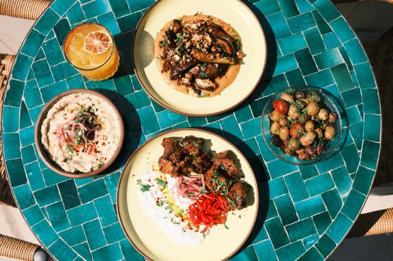Mediterranean-style appetizers on turquoise tiled table: hummus, grilled vegetables, bread, and fresh juice