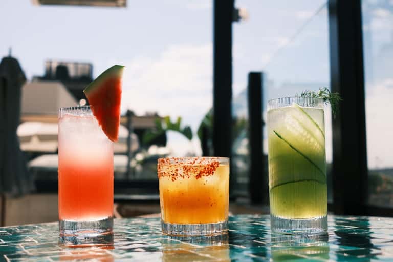 Three colorful cocktails on outdoor bar table with rooftop city views in background