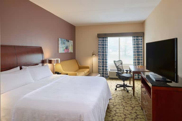 Hotel bedroom with queen bed, burgundy accent wall, sitting area with yellow chair, and patterned curtains