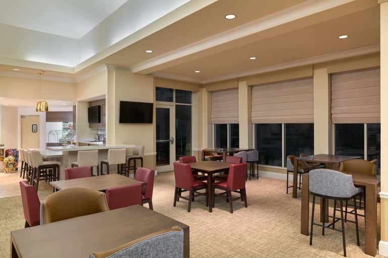 Hotel common area with multiple dining tables, burgundy chairs, bar, large windows, and bright ceiling lighting