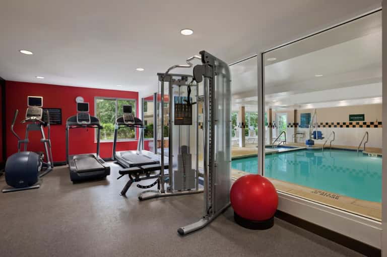 Fitness center with red wall, cardio equipment, weight machines, and glass partition overlooking indoor pool