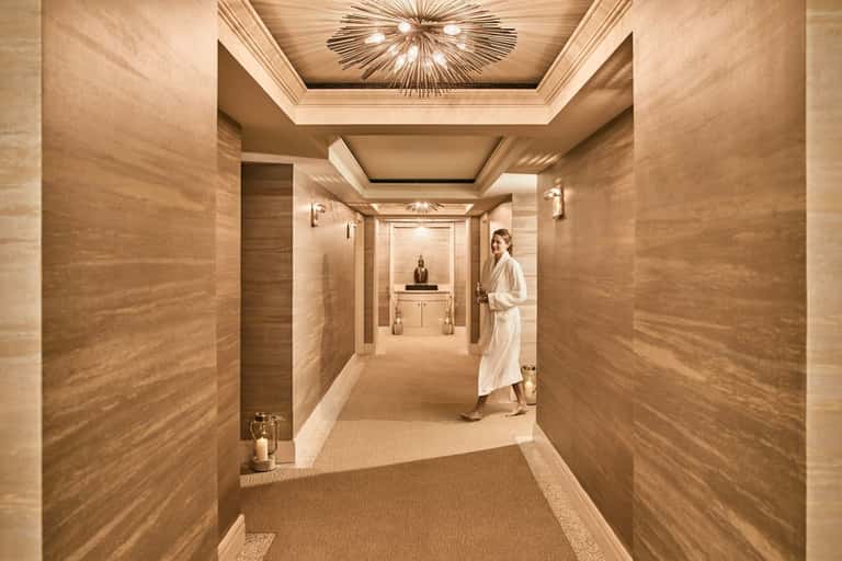 Luxury spa corridor with warm wood tones, coffered ceiling, decorative light fixture, and elegant design.
