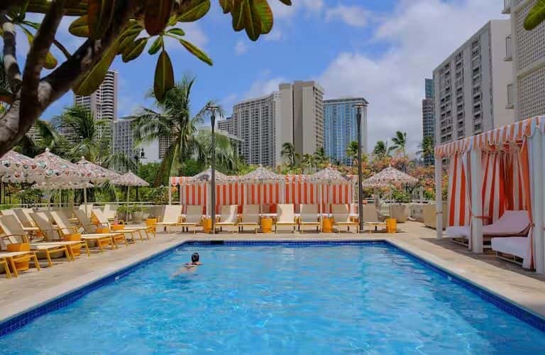 Resort pool deck with red and white striped loungers, cabanas, umbrellas, and city skyline