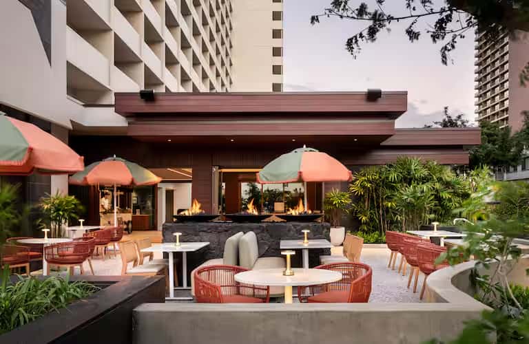 Modern outdoor patio with fireplace, coral umbrellas, woven seating, and tropical plants at dusk