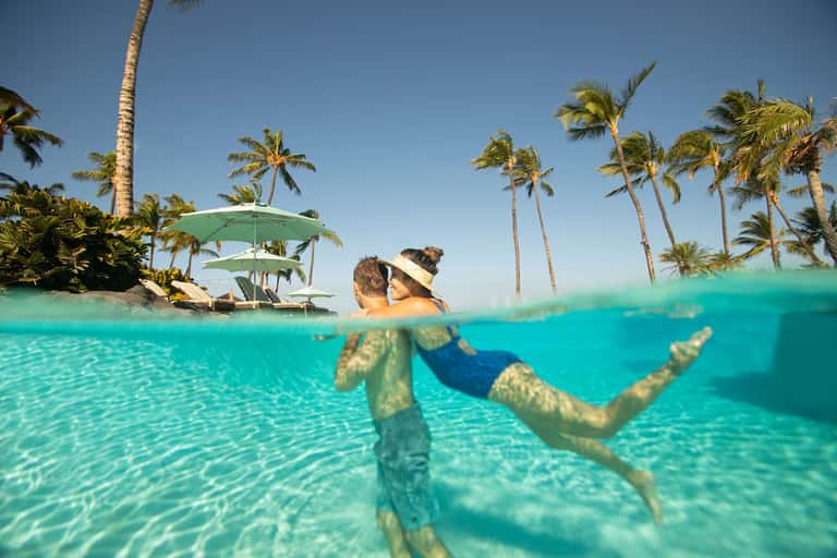 Split underwater view of two people in turquoise lagoon with palm trees and cabana on shore