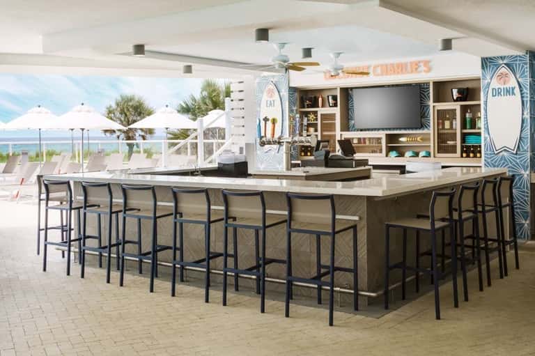 Beachfront bar and lounge area with ocean view, white umbrellas, and modern shelving displaying bottles and decor