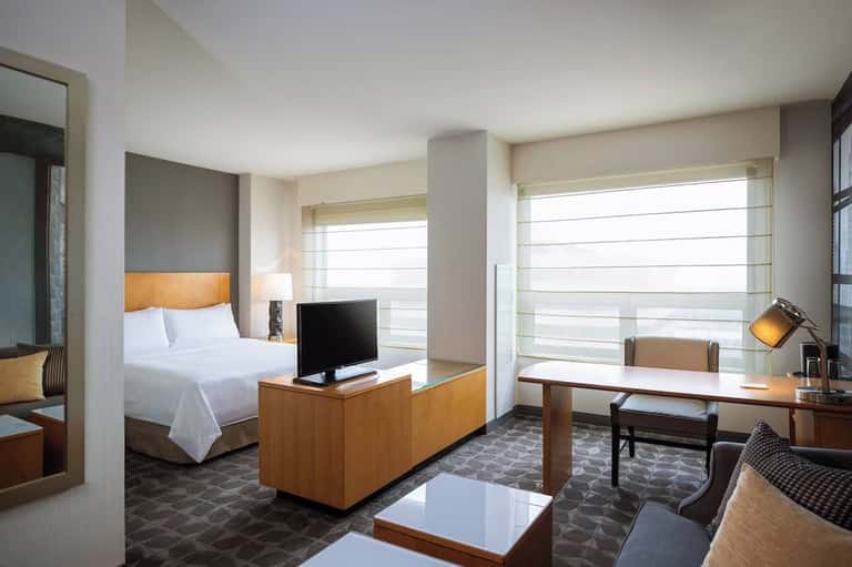 Modern hotel suite with bed, work desk, TV, and ocean view windows with sheer curtains