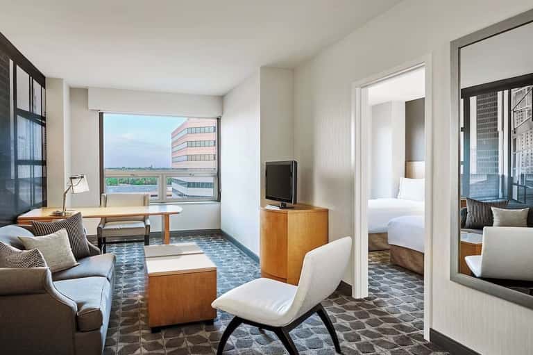 Hotel suite living area with city view, work desk, white modern furniture, and open bedroom doorway
