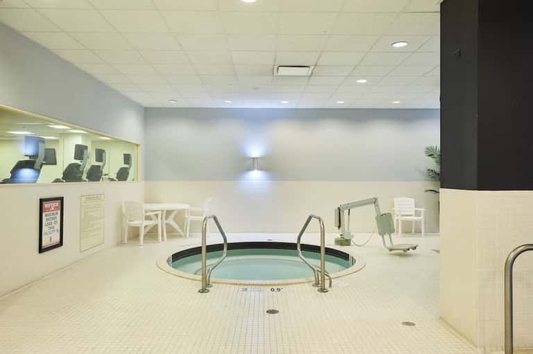Indoor hot tub with metal railings in a cream tile spa room with fitness equipment visible in background