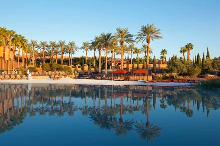 Lakeside resort with palm trees, orange umbrellas, lounge seating, and desert landscape
