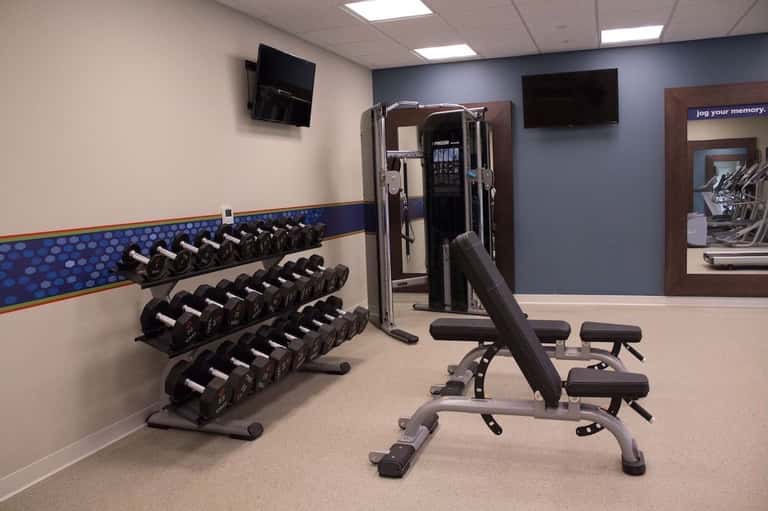 Hotel fitness center with dumbbell rack, weight bench, cable machine, and flat-screen TV
