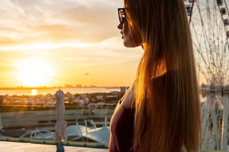 Woman with long hair at sunset viewing a Ferris wheel from a rooftop terrace overlooking water