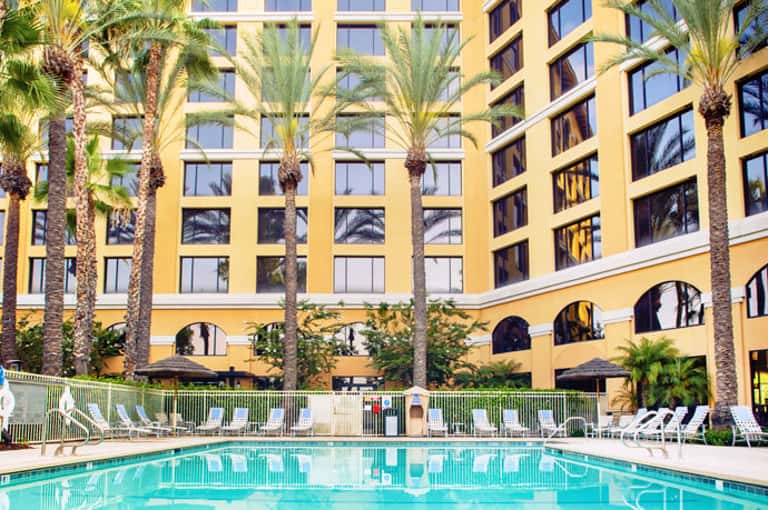 Yellow hotel building with palm trees surrounding a bright turquoise swimming pool surrounded by lounge chairs